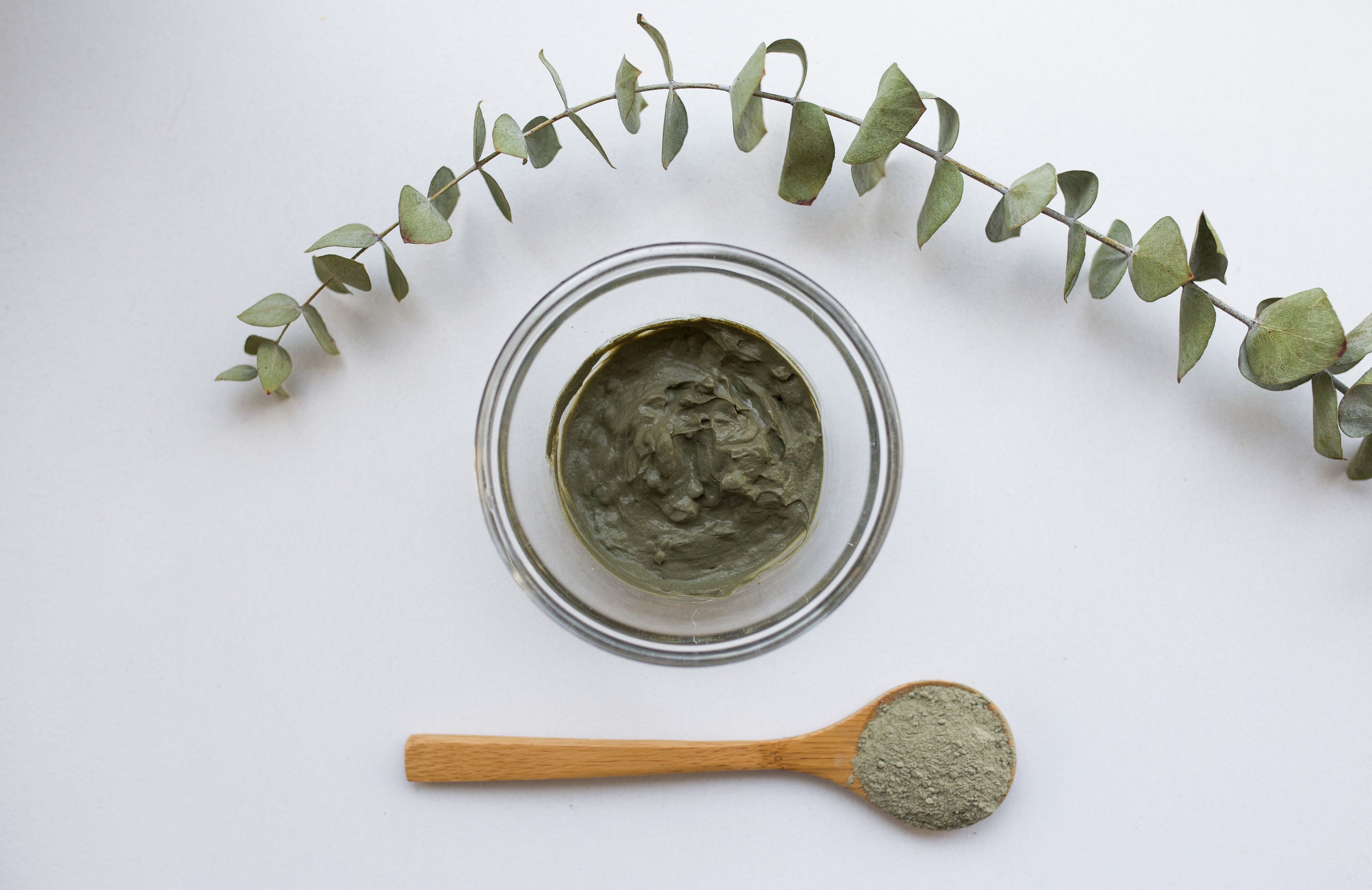 A top view of a bowl of green clay mask next to a wooden spoon with powder