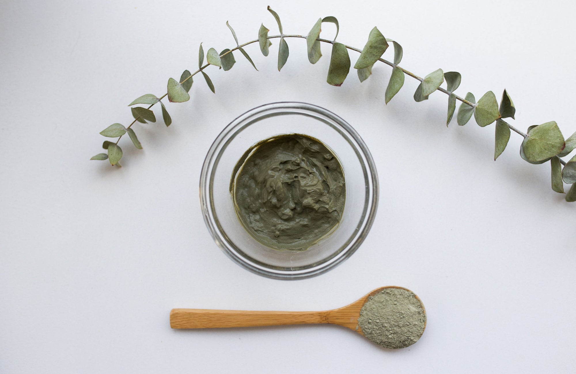A top view of a bowl of green clay mask next to a wooden spoon with powder