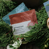 Rooibos Chai tea pouches arranged on a rustic wooden surface with green foliage and a kraft box in the background. The front pouch is labeled 'ROOIBOS CHAI' and 'RUSH HOUR', with additional teal and brown pouches visible behind it.