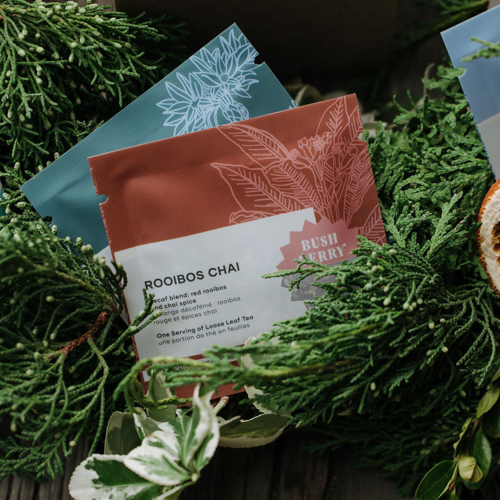 Rooibos Chai tea pouches arranged on a rustic wooden surface with green foliage and a kraft box in the background. The front pouch is labeled 'ROOIBOS CHAI' and 'RUSH HOUR', with additional teal and brown pouches visible behind it.