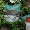 Tea product pouches arranged on a rustic wooden surface with green evergreen branches and variegated leaves around them. Visible pouches include Creamy Berry, Bush Berry, and Rooibos tea packaging.