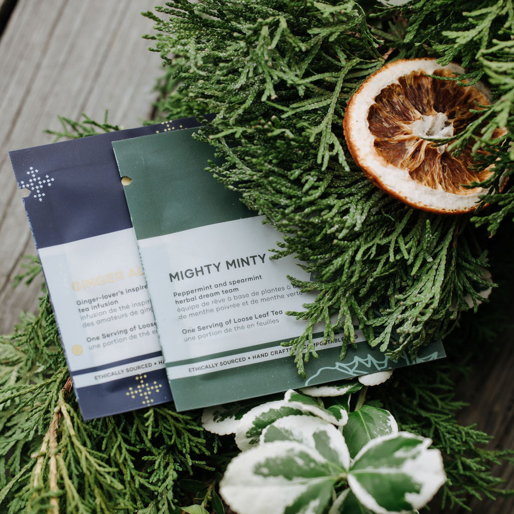 Two printed beverage cards or invitation-style inserts titled "Mighty Minty" and "Blossom" arranged on top of green evergreen branches with variegated leaves and a dried orange slice on a wooden surface.