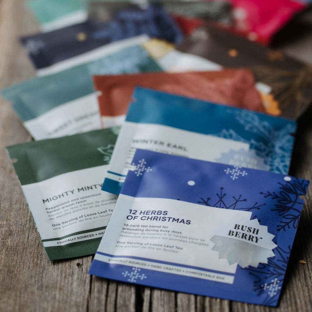 A fan of colorful herbal tea pouches arranged on a rustic wooden surface, with the front blue and white 'Rush Berry' and '12 Herbs of Christmas' pouches in sharp focus and the rest blurred in the background.