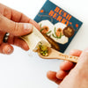 A close-up of hands preparing a small wooden spoonful of chopped green herbs and filling it into a paper wrapper, with a blue 'BUSH BERRY' box blurred in the background on a white surface.
