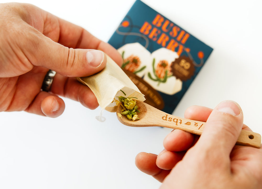 A close-up of hands preparing a small wooden spoonful of chopped green herbs and filling it into a paper wrapper, with a blue 'BUSH BERRY' box blurred in the background on a white surface.