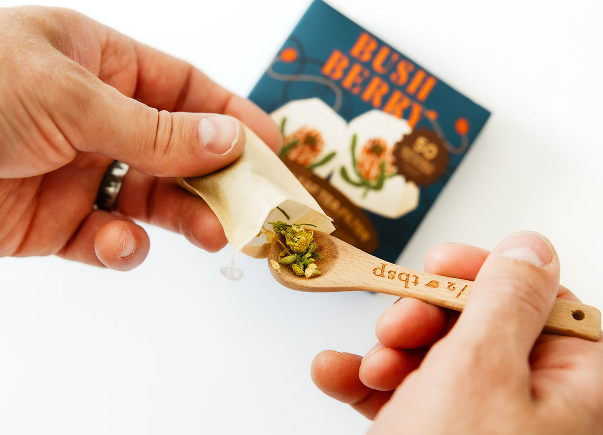 A close-up of hands preparing a small wooden spoonful of chopped green herbs and filling it into a paper wrapper, with a blue 'BUSH BERRY' box blurred in the background on a white surface.