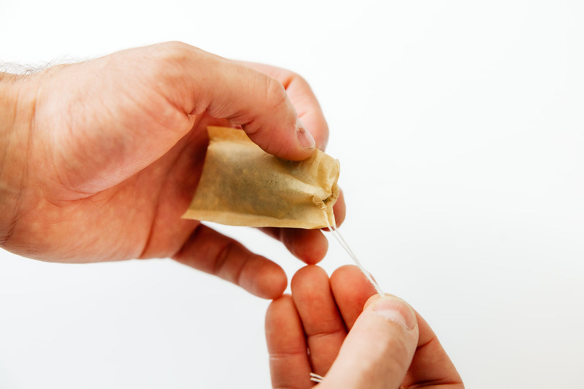 A close-up of a person's hands holding a small beige fabric pouch and pulling a thin white thread through it, against a plain white background.