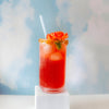 Red cocktail with a straw and floral garnish on a white pedestal against a blue sky with clouds.
