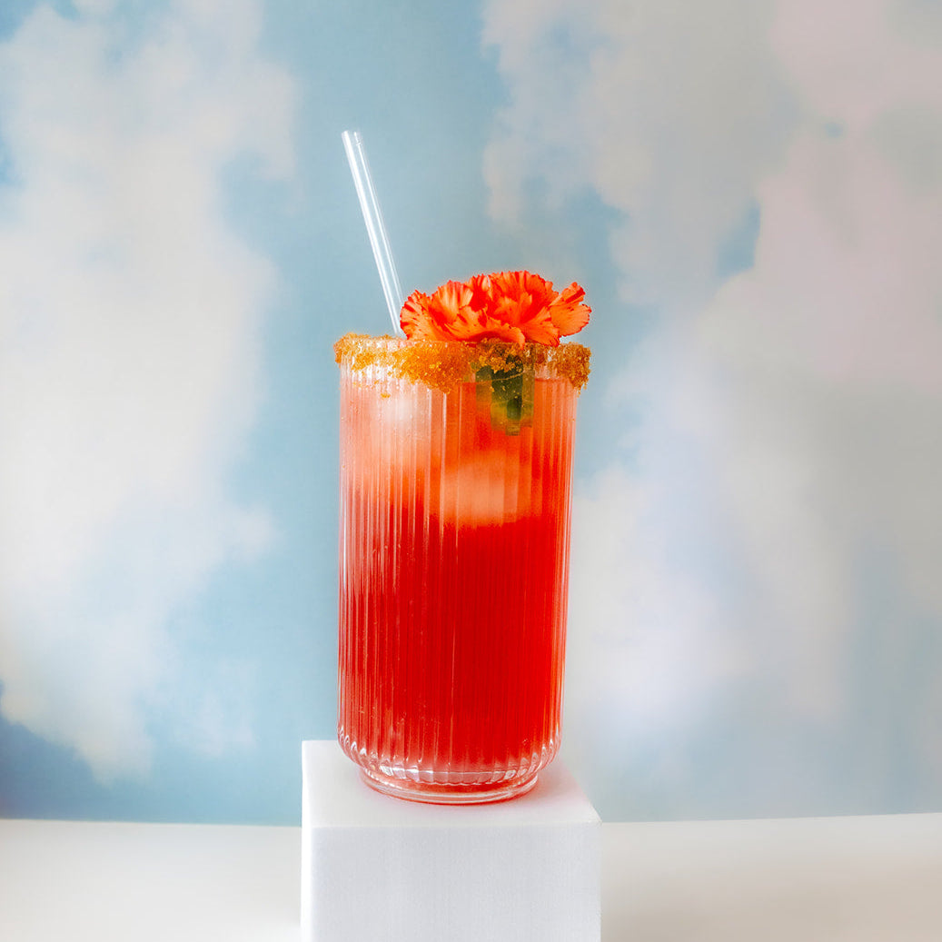 Red cocktail with a straw and floral garnish on a white pedestal against a blue sky with clouds.
