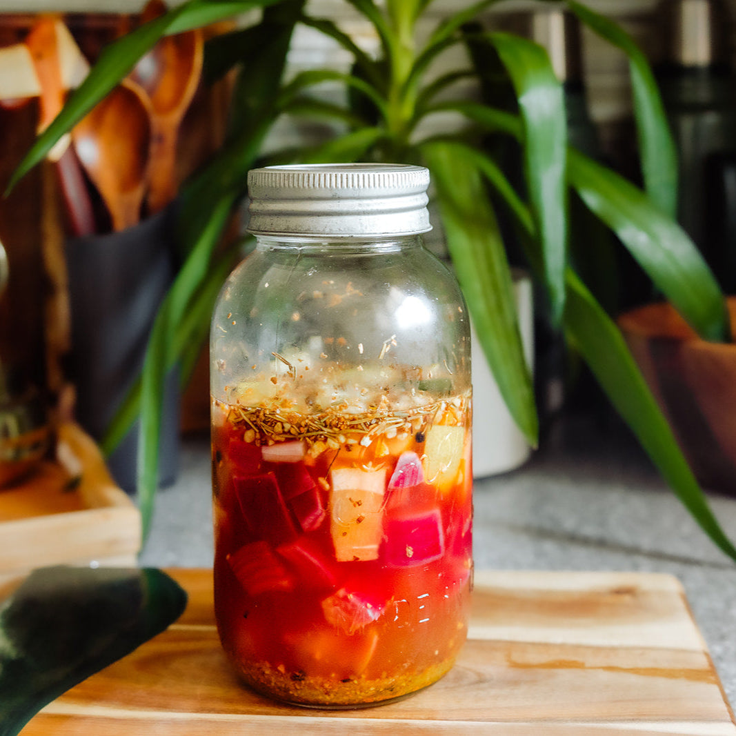 Glass jar with a metal lid containing fire cider on a wooden cutting board with plants in the background.