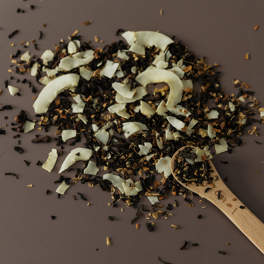 A pile of loose-leaf tea with visible white coconut flakes and dark dried botanicals, arranged on a dark brown surface with a small wooden spoon holding some of the tea.