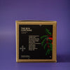 A square kraft paper box with a black front label reading 'THE BOX CONTAINS' and holiday greenery graphics, photographed on a purple background.