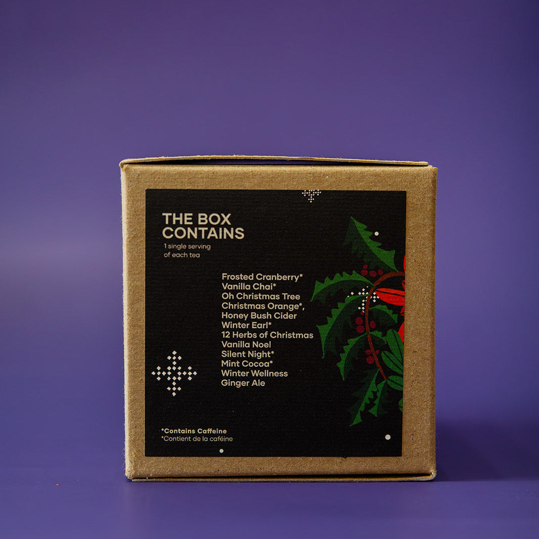 A square kraft paper box with a black front label reading 'THE BOX CONTAINS' and holiday greenery graphics, photographed on a purple background.